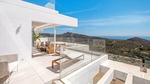 A modern outdoor terrace with a glass balustrade, furnished seating, and a scenic view of the landscape. - Palo Alto | Marbella Luxury Rooftop Penthouse (Ojén)