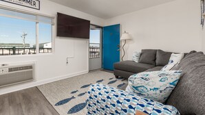 Una sala de estar con un sofá gris, una puerta azul, un televisor de pantalla plana montado en la pared y una ventana con persianas.