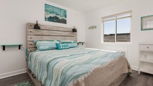 Un dormitorio con cabecera de madera, colcha a rayas, almohada con temática de playa, ventana con vista al mar y una pequeña estantería con un distribuidor de corriente.