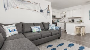 Una sala de estar moderna con un sofá seccional gris, almohadones decorativos, una obra de arte de red en la pared y una cocina al fondo.