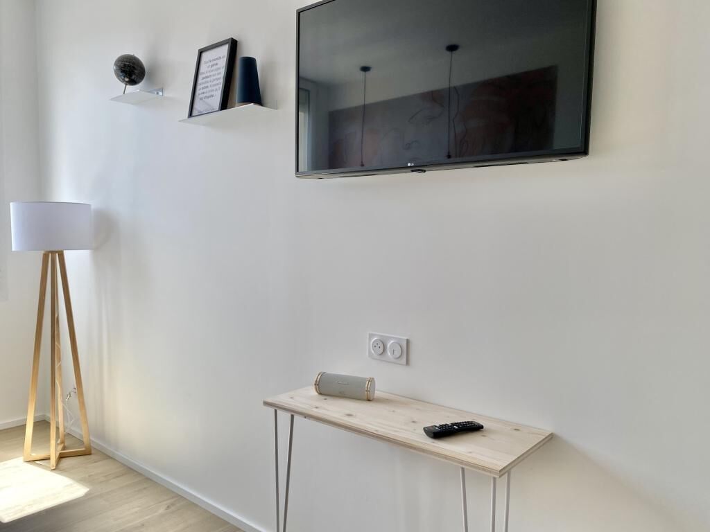 A modern room with a flat-screen TV mounted on the wall, a small wooden table with a remote control, and a floor lamp.