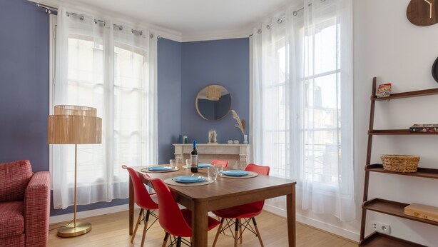A dining area with a wooden table set for four, red chairs, a floor lamp, and a bookshelf.