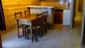 Un intérieur en bois comprenant une table et des chaises, un coin cuisine avec des meubles blancs et une porte-fenêtre donnant sur une autre pièce.