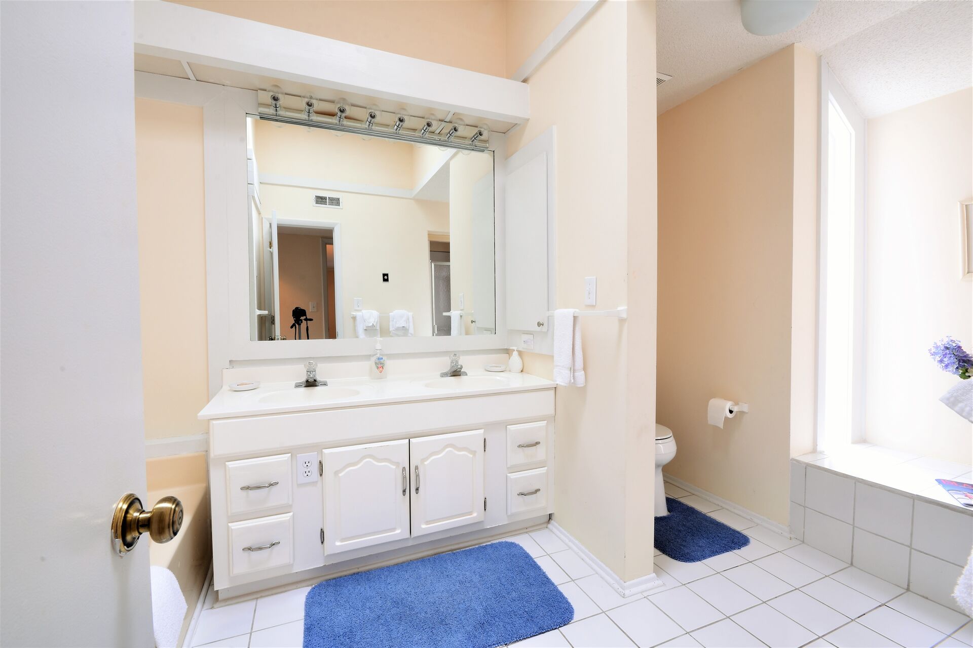 A bathroom with a white sink, mirror, and cabinets, a toilet, and a blue bath mat.