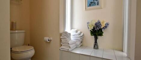A bathroom with a toilet, a roll of toilet paper, a stack of towels, and a vase with flowers.