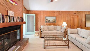 A living room with a fireplace, wooden walls, a ceiling fan, a sofa, a glass coffee table, and a bookshelf. - Longs for the Sea - North Litchfield Beach (Pawleys Island)