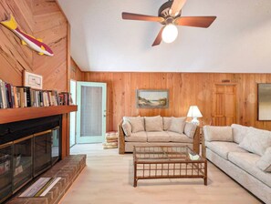 A cozy living room with a fireplace, wooden walls, a ceiling fan, and a bookshelf.