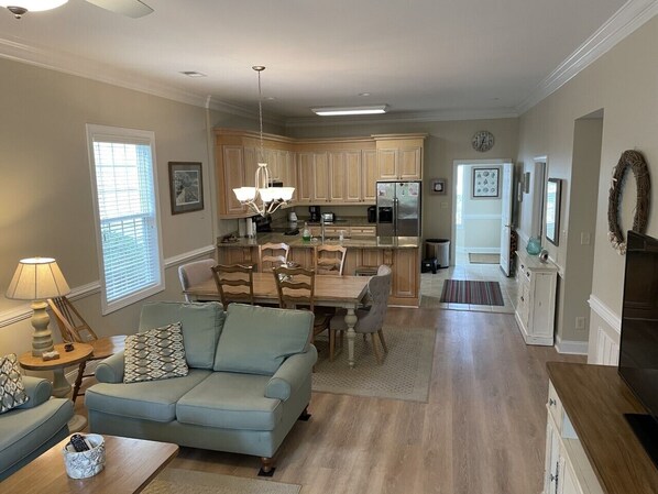A spacious living room with a light blue sofa, a dining table with chairs, and a kitchen area with wooden cabinets and a refrigerator.