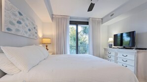 Un dormitorio con cama, ventilador de techo, televisor y ventana con cortinas.