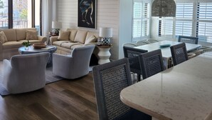 Una sala de estar moderna con un conjunto de sofás, un comedor con mesa y sillas, y un amplio ventanal con persianas.