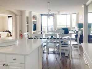 A bright, spacious kitchen with white cabinetry, a dining area with white chairs, and a large window providing natural light. - Crescent D3D  Oceanfront Condo - Litchfield by the Sea - Community Pool & Hot Tu (Pawleys Island)