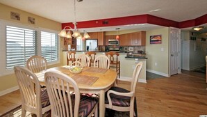 A spacious dining area with a wooden table and chairs, a kitchen in the background, and a red accent wall. - Oceanfront Condo - Litchfield by the Sea - Community Pool & Hot Tub (Pawleys Island)