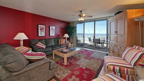 A living room with a red wall, a ceiling fan, a sofa, a coffee table, and a view of the balcony. - Oceanfront Condo - Litchfield by the Sea - Community Pool & Hot Tub (Pawleys Island)