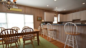 A dining area with wooden chairs and a table, a kitchen with white cabinets, and a green rug on the floor. - Beautiful Ames to Please (Pawleys Island)