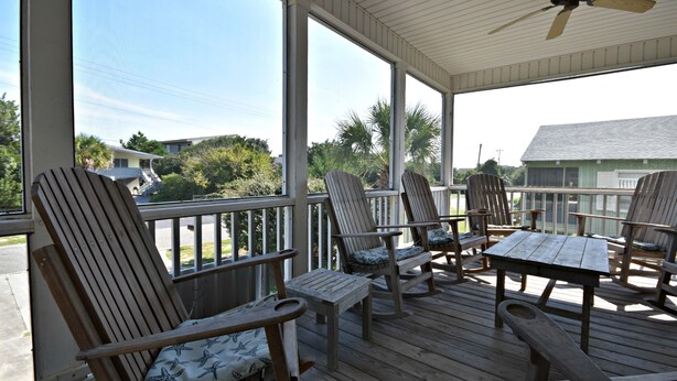 A covered outdoor deck with wooden chairs and tables, large windows, and a ceiling fan. - Beautiful Ames to Please (Pawleys Island)