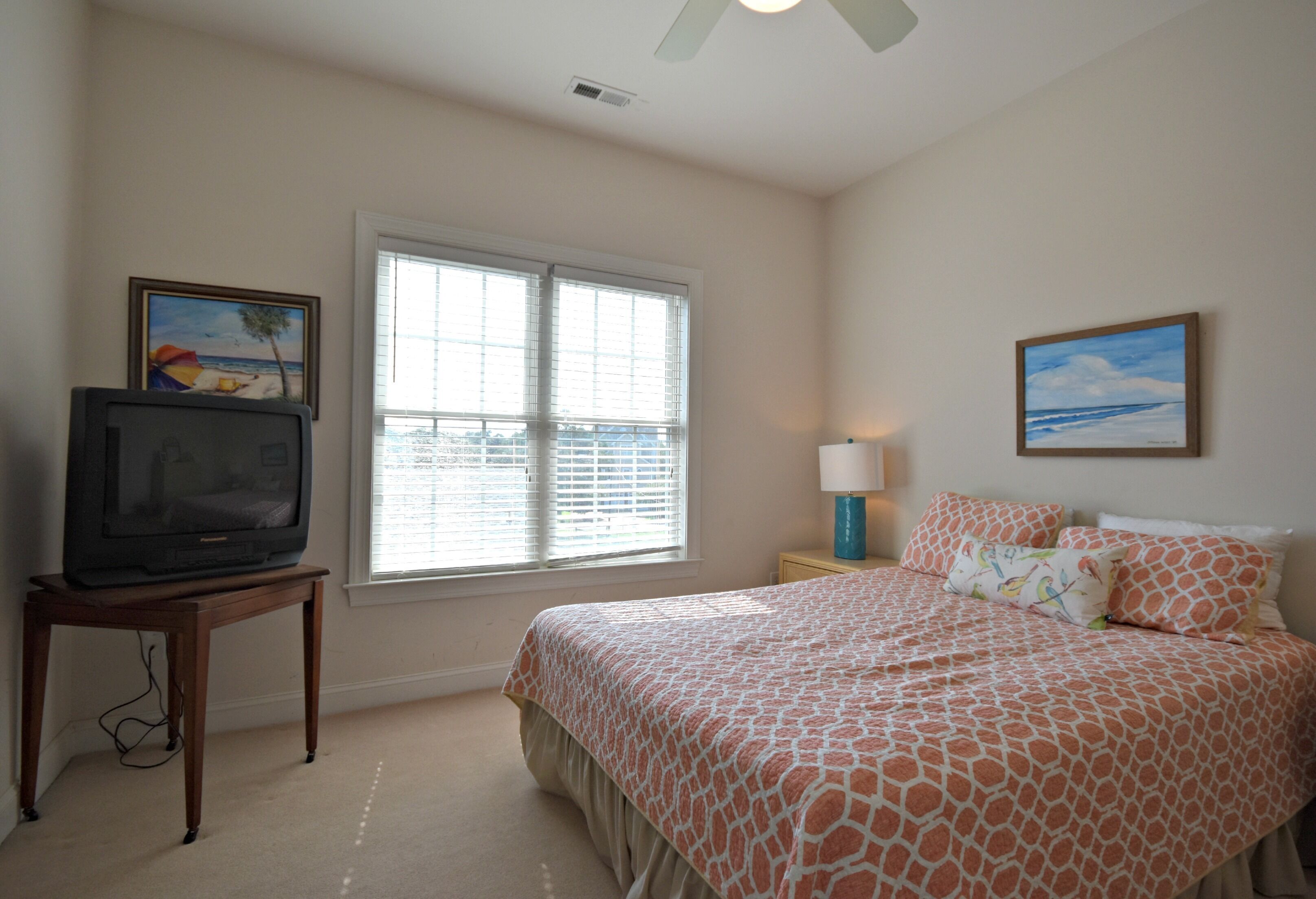 A bedroom with a bed, a television on a stand, a window with blinds, and two paintings on the walls.