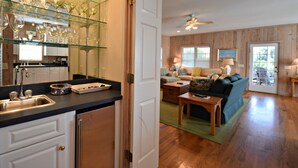 A kitchen with a glass wine rack, a sink, and a countertop. There is a dining area with a sofa and a table. - Beautiful Ames to Please (Pawleys Island)