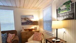 Un salón acogedor con un sofá marrón, una butaca, una mesita de madera y una lámpara. Un cuadro de una playa adorna la pared, y se ve un cartel que dice "MOMA'S BEACH HOUSE".