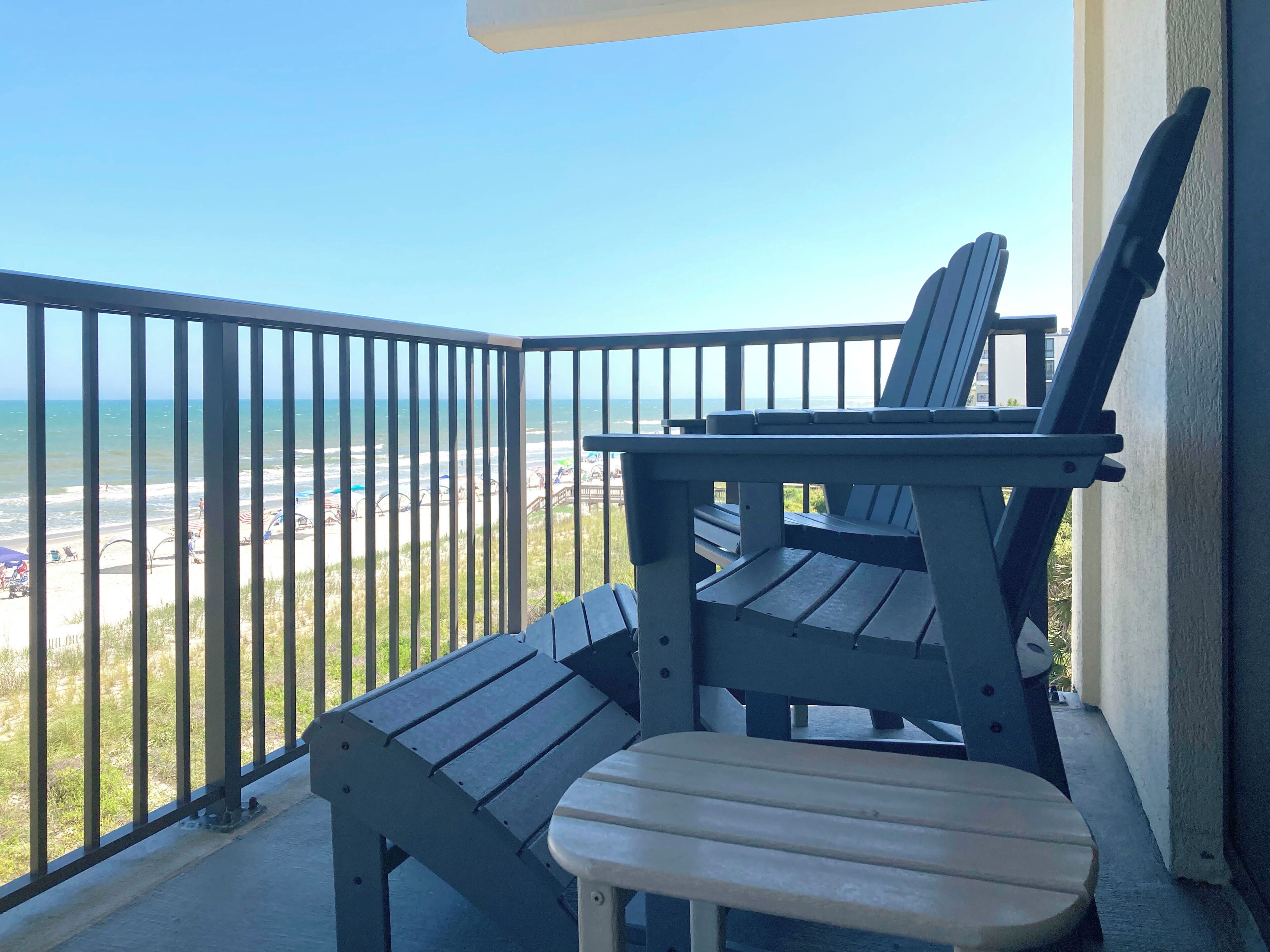A balcony with a view of the beach, a table, and two chairs.