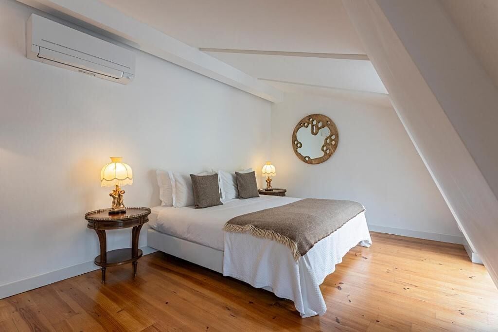 Quarto com cama, duas mesas de cabeceira com abajures, um espelho fixado na parede e piso de madeira.