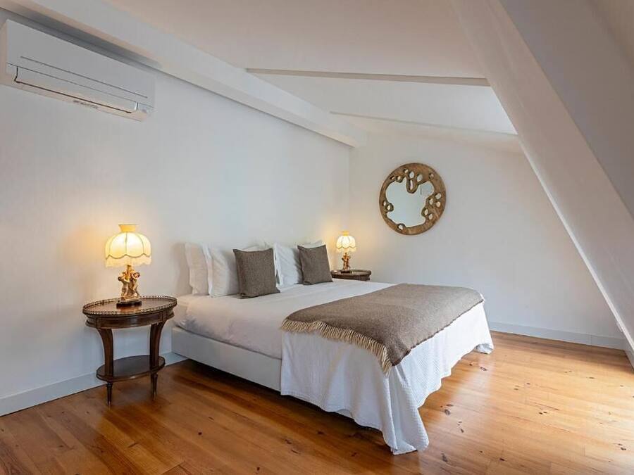 Quarto com cama, duas mesas de cabeceira com abajures, um espelho fixado na parede e piso de madeira.