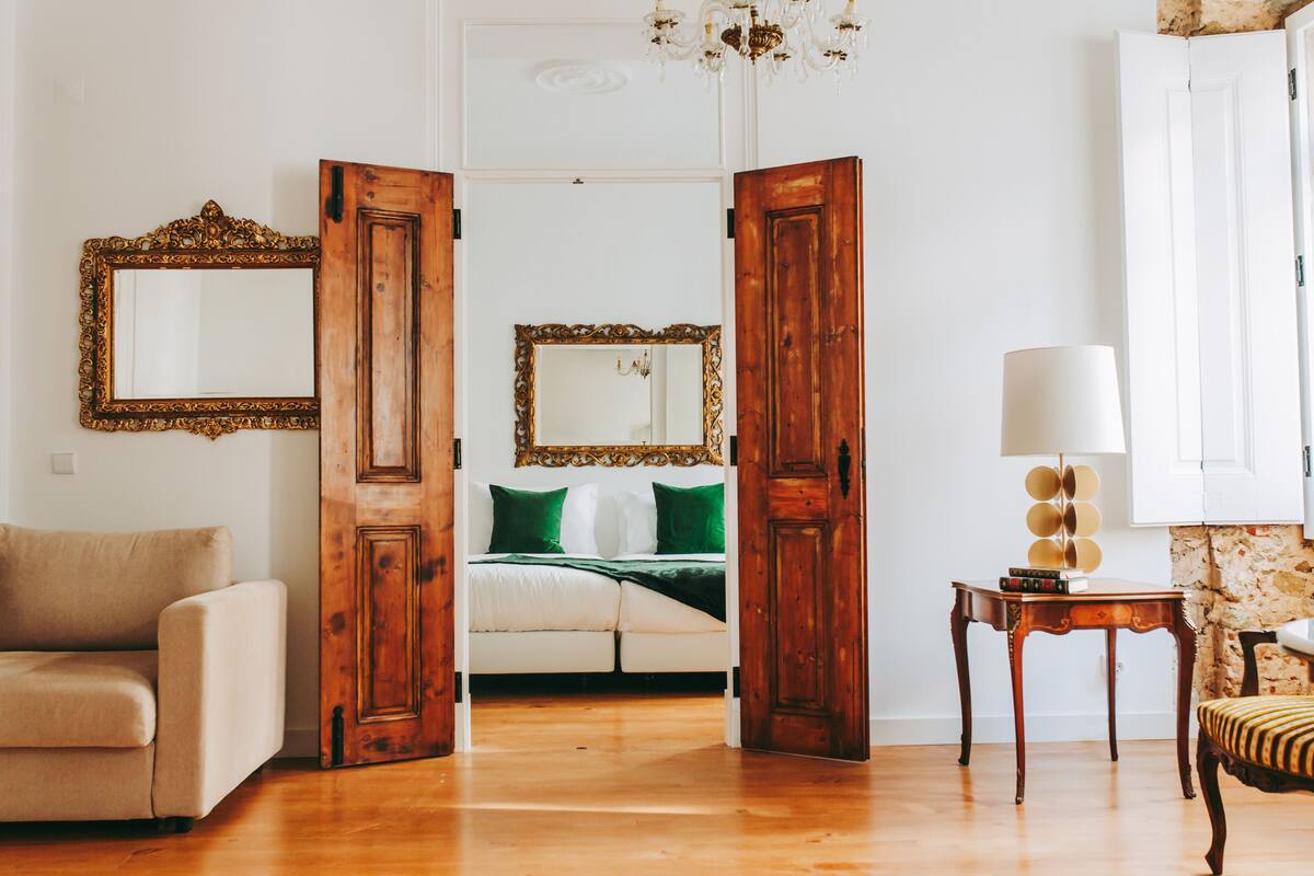 A room with a wooden door, a bed with green pillows, a white sofa, and a small table with a lamp.