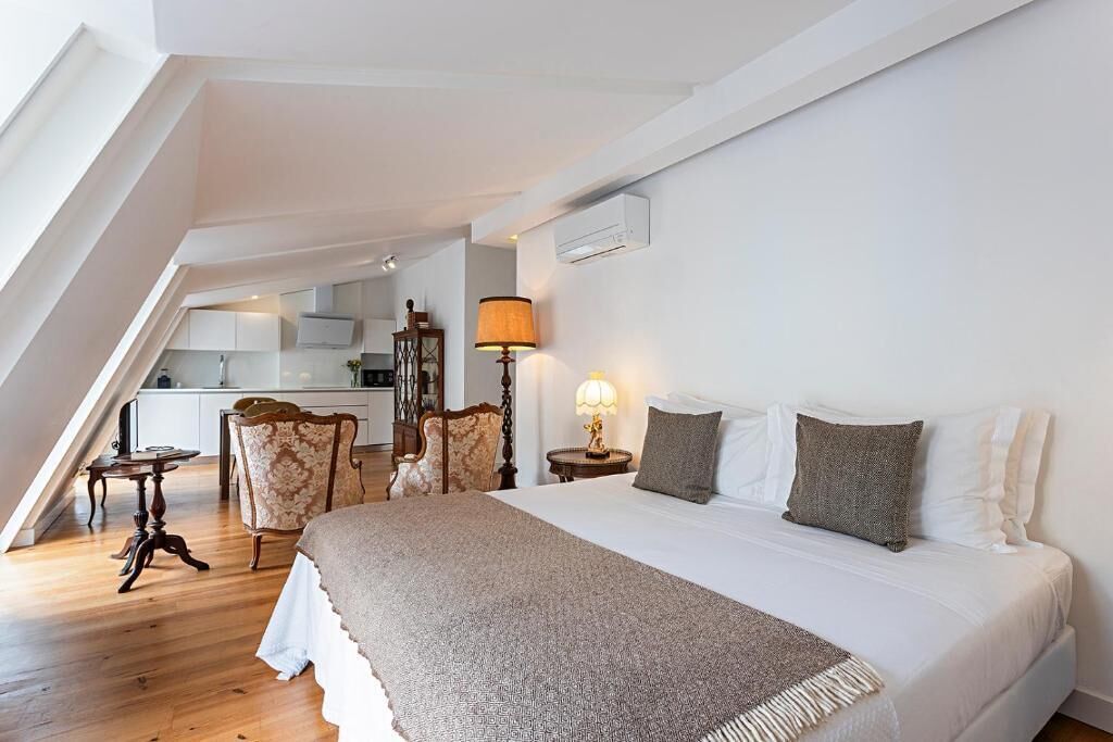 Ein Schlafzimmer mit einem großen Bett, einem Schreibtisch und einem Stuhl. Im Hintergrund ist eine Küche zu sehen.