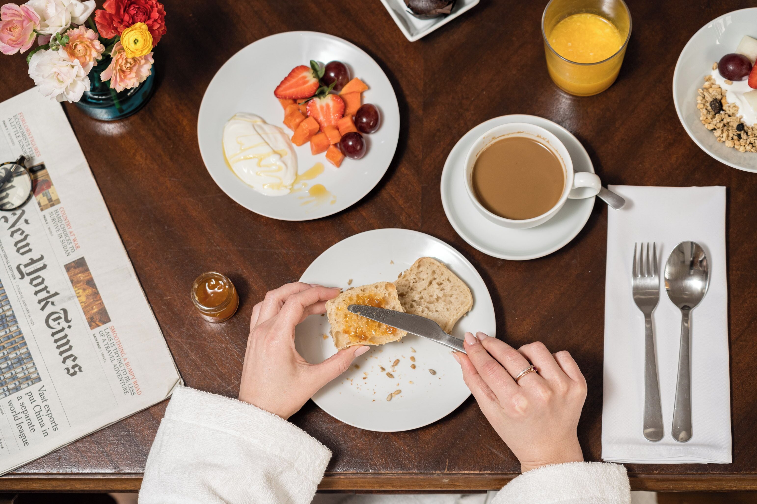 Una mesa de desayuno con periódico, un plato de fruta, un vaso de jugo de naranja, una taza de café y cubiertos.