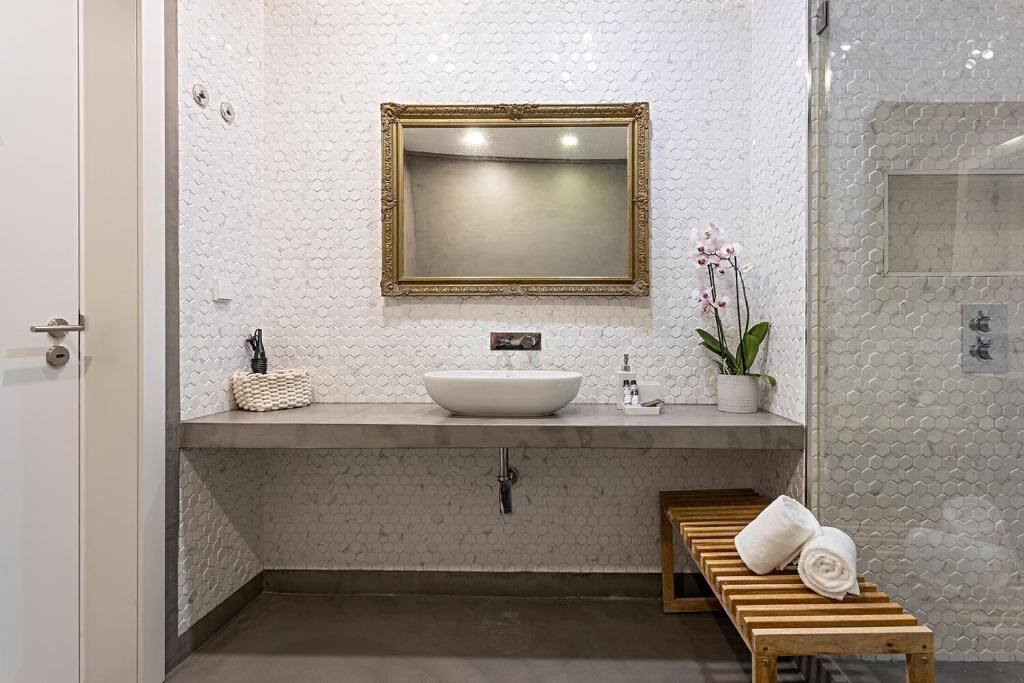 A bathroom with a white sink, a gold-framed mirror, a wooden bench with towels, and a potted orchid.