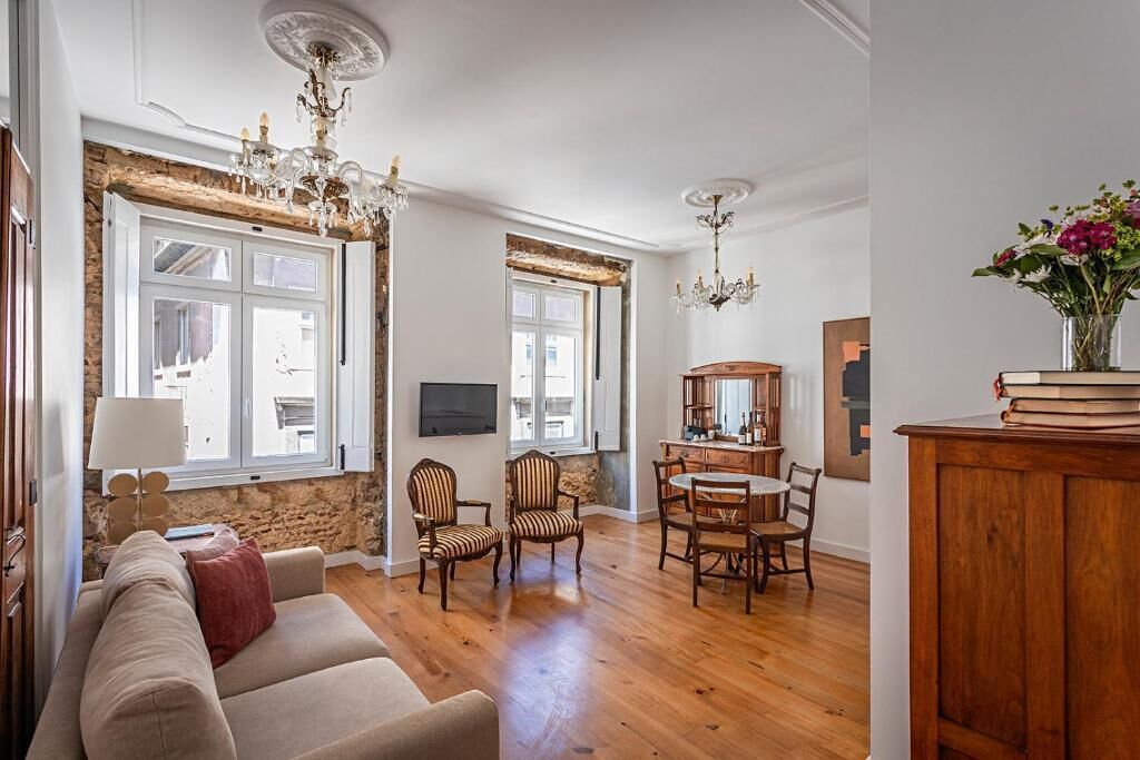 A spacious living room with a wooden floor, a beige sofa, striped armchairs, a dining table with chairs, and a chandelier.