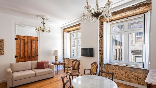 A living room with a wooden floor, a round table, and a sofa. There are two chairs, a TV, and a chandelier. - 54 São Paulo – Exclusive Apartment Hotel (Lisbon)