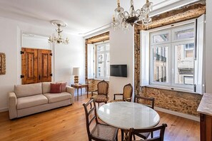 A living room with a wooden floor, a round table, and a sofa. There are two chairs, a TV, and a chandelier. - 54 São Paulo – Exclusive Apartment Hotel (Lisbon)