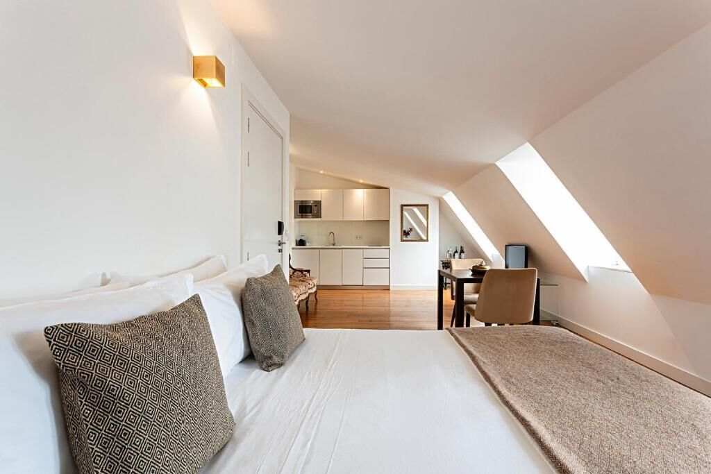 Un dormitorio con cama, almohadas, un escritorio y una kitchenette al fondo.