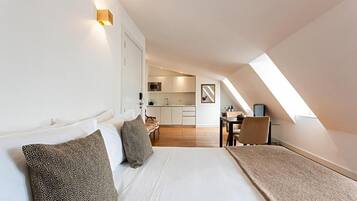 Un dormitorio con cama, almohadas, un escritorio y una kitchenette al fondo.