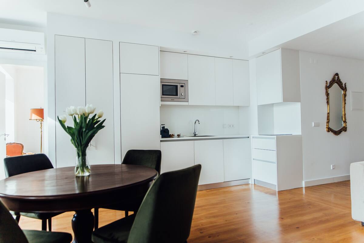 A modern kitchen with white cabinets, a microwave, and a sink. There is a dining table with chairs and a vase of flowers.