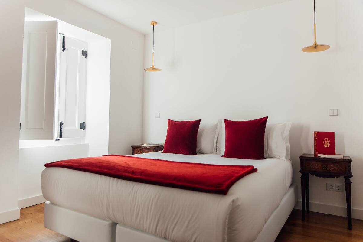 A bedroom with a bed, red pillows, a red throw blanket, a nightstand with a book, and a window with curtains.