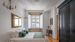 A bedroom with a bed, a chandelier, a window, a mirror, and a wardrobe. - 54 São Paulo – Exclusive Apartment Hotel (Lisbon)