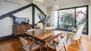 A dining area with a wooden table, four chairs, a vase with flowers, a television on a cabinet, and a large window with a view of trees and flowers. - 54 São Paulo – Exclusive Apartment Hotel (Lisbon)