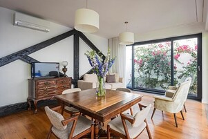 Un comedor con mesa de madera, cuatro sillas, un jarrón con flores, un televisor sobre un mueble bajo y un ventanal con vistas a árboles y flores.