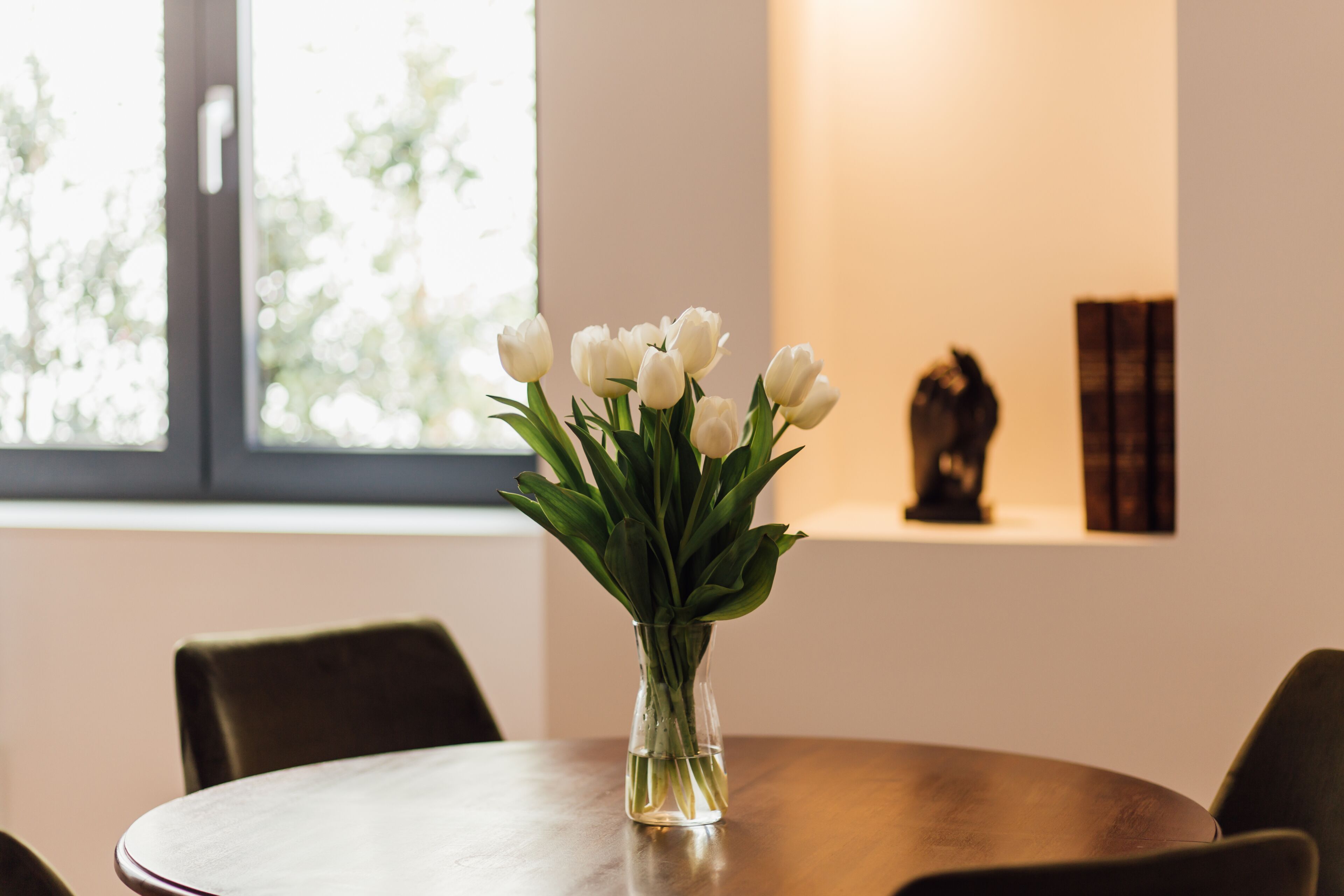 Una mesa redonda de madera con un florero de vidrio transparente que contiene tulipanes blancos.
