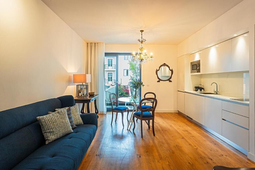 A modern living room with a dark blue sofa, a dining table with chairs, a chandelier, and a kitchen area with white cabinets and a microwave.