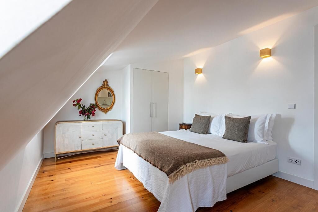 Een slaapkamer met een schuine kap, een bed met witte beddengoed en twee kussens, een witte dressoir met spiegel, en een vaas met bloemen.