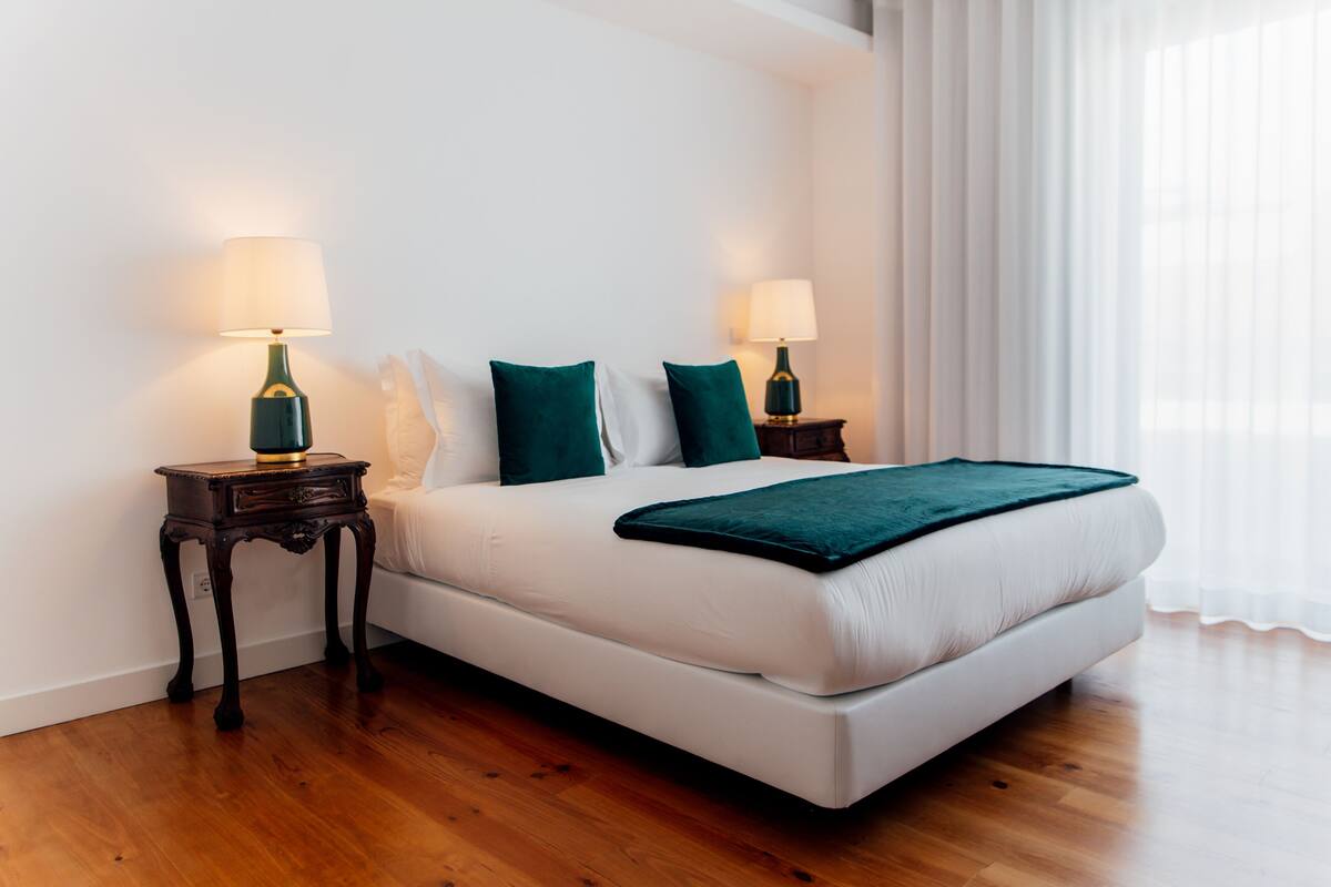 A neatly made bed with white linens and two green pillows, flanked by two bedside tables with lamps, in a room with wooden flooring and white walls.