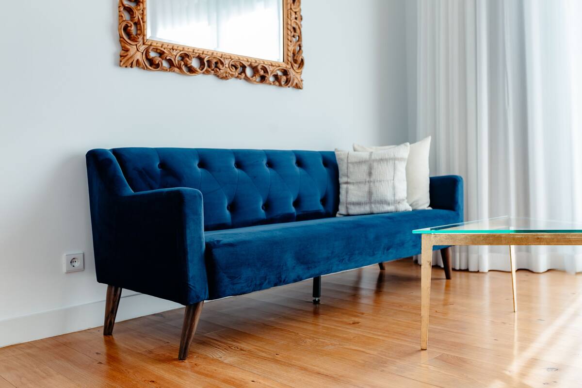 A blue velvet sofa with wooden legs, a glass coffee table, and a decorative mirror on the wall.