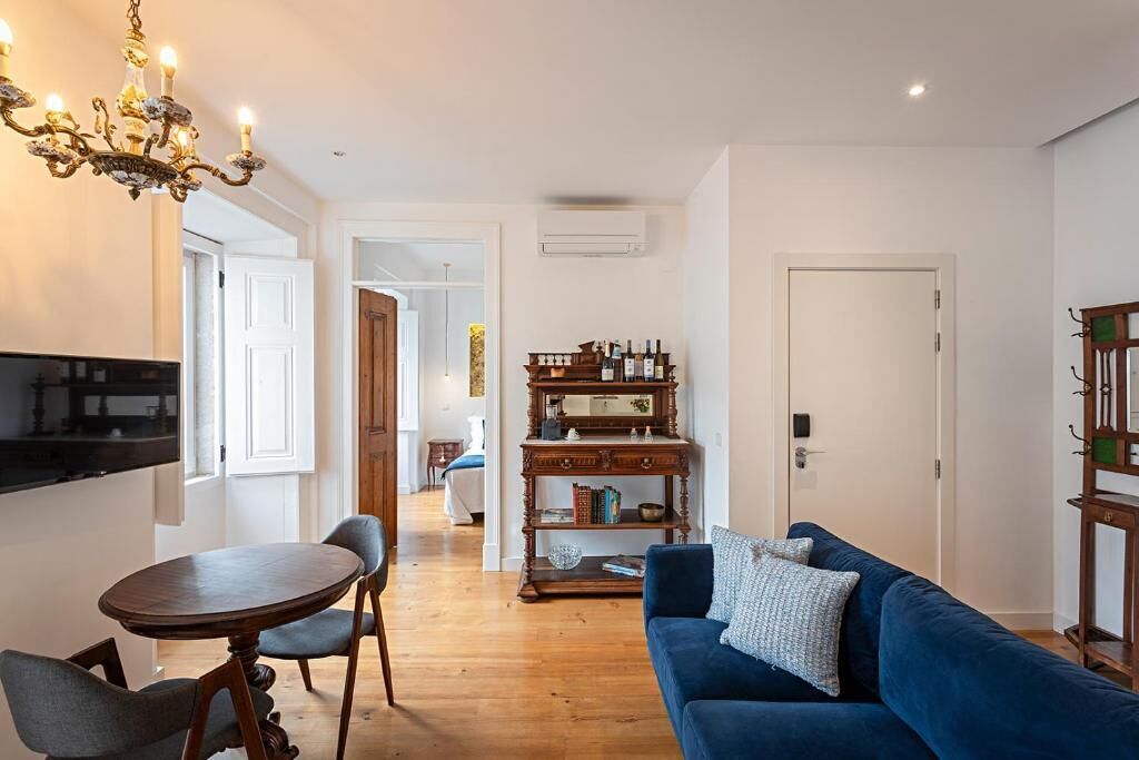 A modern living room with a blue sofa, a wooden coffee table, and a chandelier. There is a bookshelf with books and decorative items, and a door leading to another room.