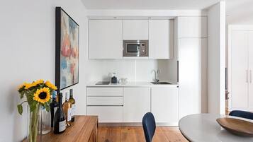 Uma cozinha moderna com armários brancos, micro-ondas embutido e uma pia. Há uma área de jantar com mesa e cadeiras de madeira, e um vaso com girassóis na mesa.