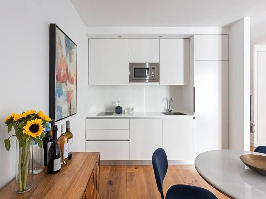 Uma cozinha moderna com armários brancos, micro-ondas embutido e uma pia. Há uma área de jantar com mesa e cadeiras de madeira, e um vaso com girassóis na mesa.