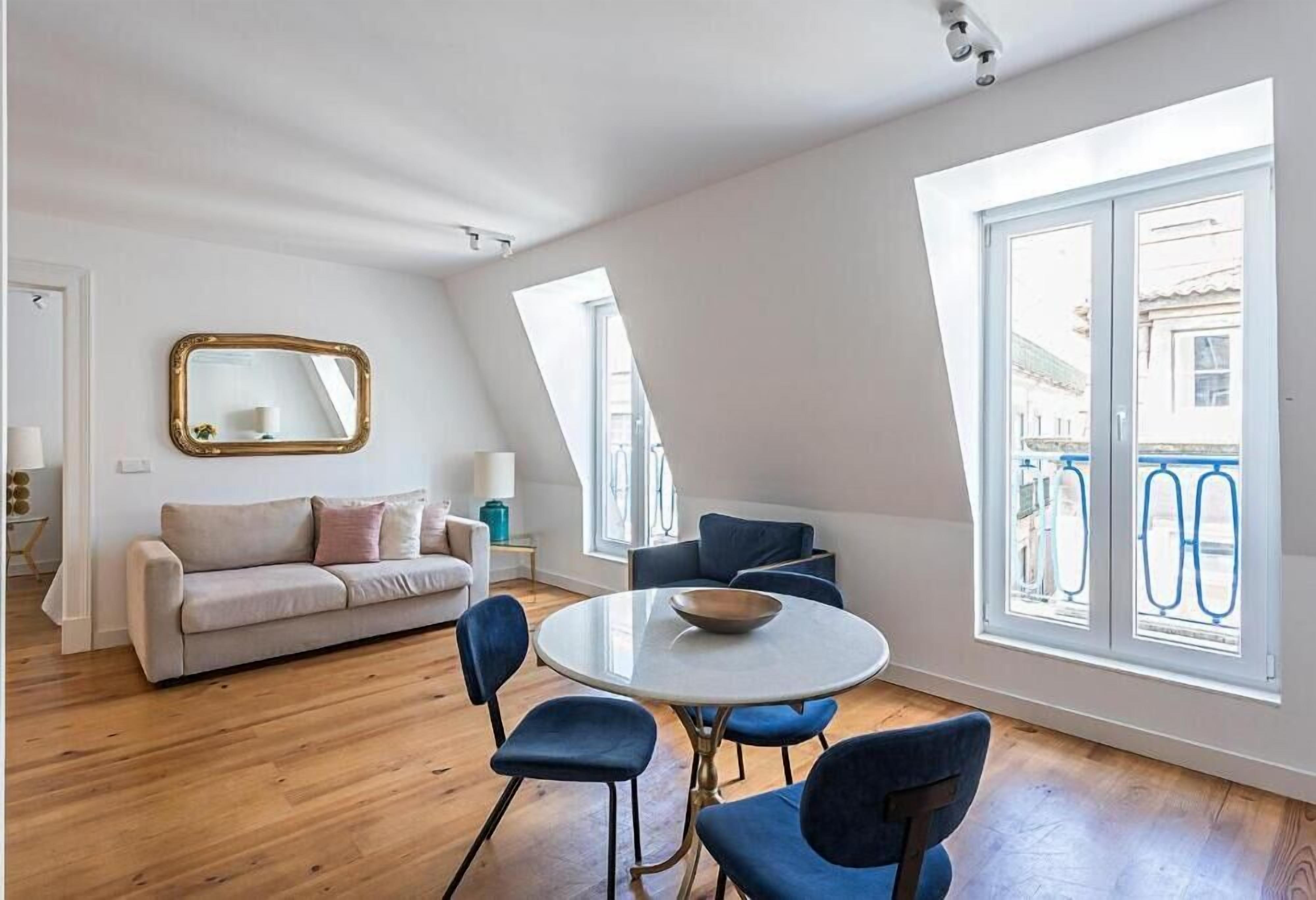 A modern living room with a round table, blue chairs, a beige sofa, and a large window with a balcony view.