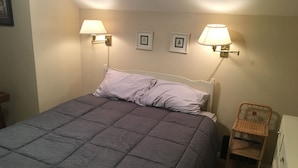 Ein ordentlich bezogenes Bett mit einer grauen Steudecke, zwei an der Wand befestigten Lampen und ein Korbstuhl neben einem Schminktisch.