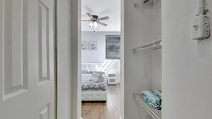 Un pasillo estrecho que conduce a un dormitorio con cama, ventilador de techo y ventana con persianas.